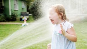 Little girl with facing a spraying hose with her mouth open. She is soaking wet.