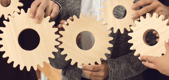 Series of wooden gears in a horizontal line, each held by a person's fingers.