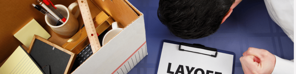 A box with office supplies next to a clipboard with a sheet of paper that says "layoff" on it and a man with his head and fist on the desk indicating devastation.
