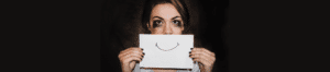 Close up of a white woman's face with her mascara smeared to the sides of her eyes, holding a white piece of paper in front of her with a line drawing of a smiley mouth. The effect is a woman who is very unhappy but faking a smile for others.