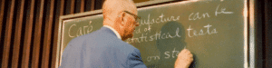 Dr. Deming in his 80s, standing with his back to the audience writing on a green chalkboard. He is wearing a blue sports coat, has a bald head, and wears glasses.