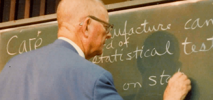 Dr. Deming in his 80s, standing with his back to the audience writing on a green chalkboard. He is wearing a blue sports coat, has a bald head, and wears glasses.